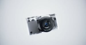 A compact digital camera with a transparent body, revealing internal components, is centered against a plain light gray background.