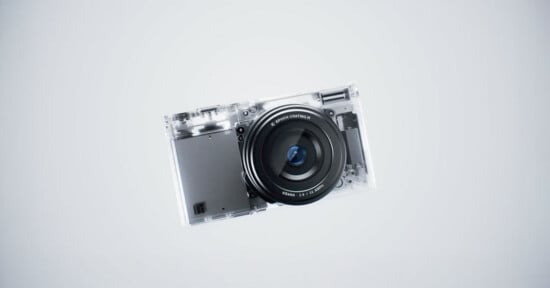 A compact digital camera with a transparent body, revealing internal components, is centered against a plain light gray background.