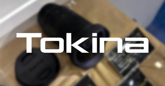 A blurred photo of a camera lens and lens cap on a table, with the word "Tokina" in bold white letters prominently overlaid in the center.