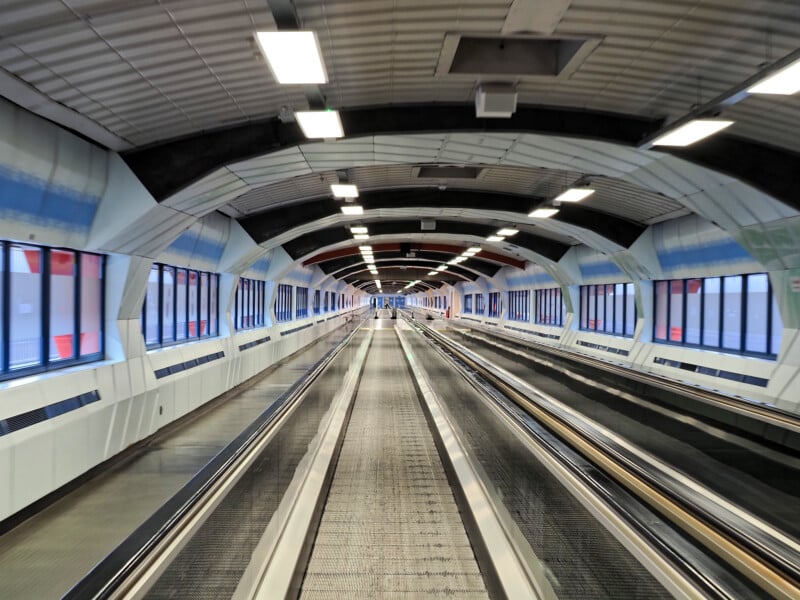 A modern, empty airport walkway with metal moving walkways, enclosed by a tunnel-like structure with curved ceiling, windows on both sides, and bright overhead lights.