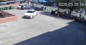 A white car drives through a mostly empty parking lot on a sunny day. Several parked cars and green trash bins are visible in the background. The timestamp on the image reads "2026-03-26 12:48.