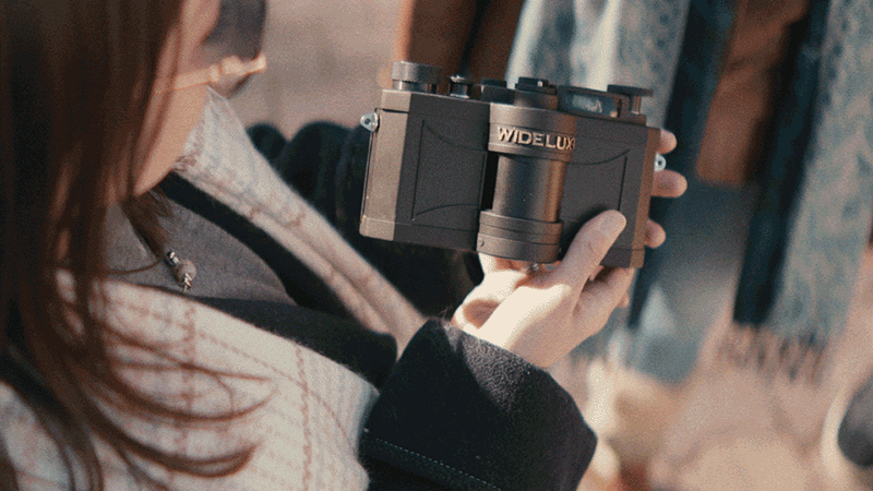 A person wearing glasses and a patterned coat holds a vintage Widelux panoramic camera, looking down at it while standing outdoors.