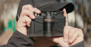 A person wearing a black cap holds a Widelux panoramic film camera up to their face, preparing to take a photo outdoors.