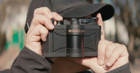 A person wearing a black cap holds a Widelux panoramic film camera up to their face, preparing to take a photo outdoors.