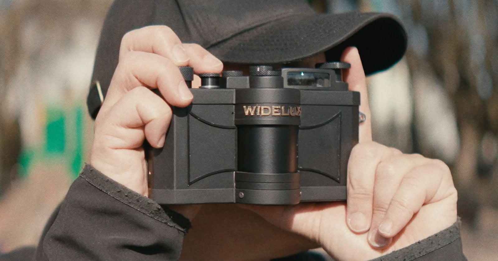 A person wearing a black cap holds a Widelux panoramic film camera up to their face, preparing to take a photo outdoors.