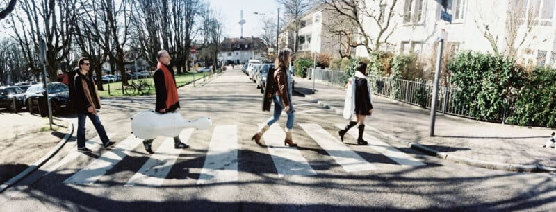 Four people walk across a zebra crossing on a sunny street, one carrying a white instrument case, mimicking the iconic Abbey Road album cover by The Beatles. Trees and buildings line the road in the background.