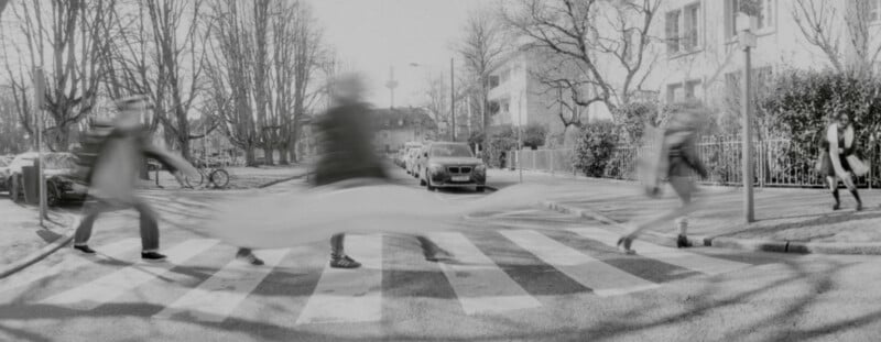 A blurred black-and-white photo shows people hurriedly crossing a zebra-striped crosswalk on a city street lined with leafless trees and parked cars on a sunny day.