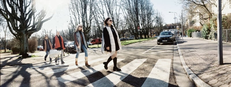 Four people cross a sunny street at a pedestrian crosswalk, bundled in winter clothing. Leafless trees line both sides, and parked cars are visible in the background. The scene echoes the famous Abbey Road photo.