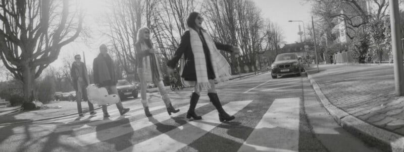 Four people cross a street at a crosswalk on a sunny, tree-lined road, with a car waiting nearby. The scene resembles the iconic Abbey Road album cover. The image is in black and white.