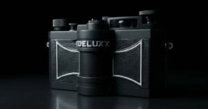 A black vintage-style camera labeled “DELUX” is shown against a dark background, with textured panels and metallic knobs visible in dramatic lighting.