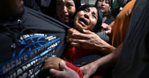 A distressed woman is held tightly by several people, her face showing anguish as she cries out. The scene appears chaotic and emotional, with hands gripping her and tense expressions on surrounding faces.