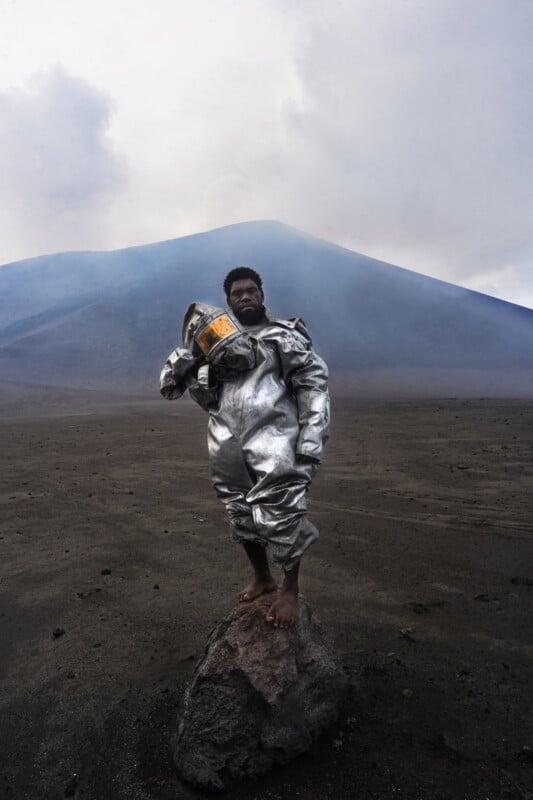 A person in a shiny silver protective suit stands barefoot on a rock in a barren, dark volcanic landscape, holding a helmet, with a misty mountain and cloudy sky in the background.