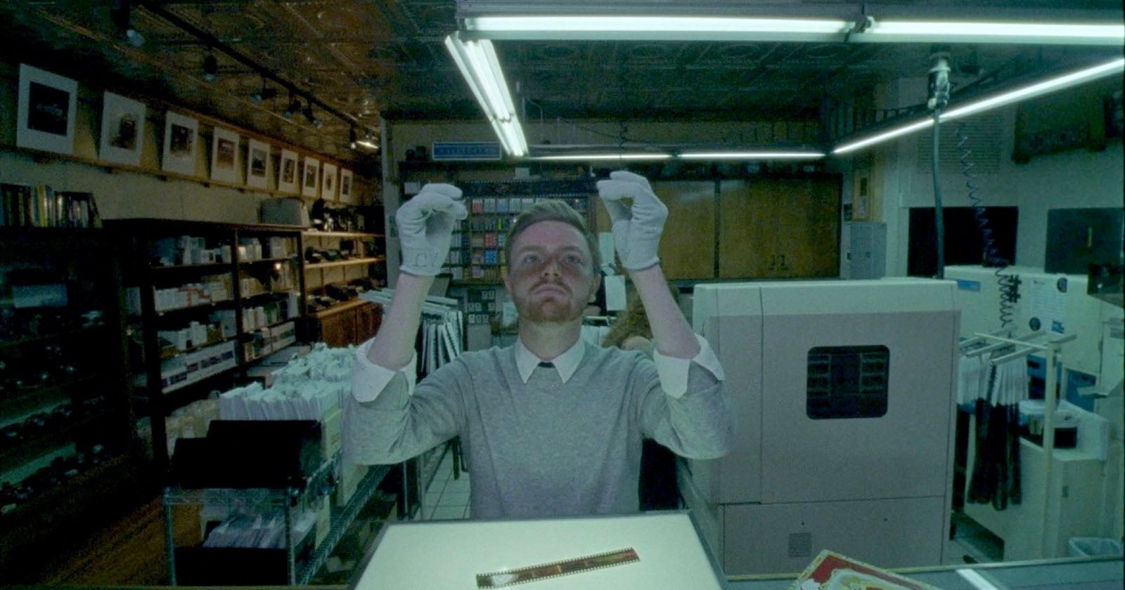 A man wearing white gloves examines photographic film in a dimly lit photo lab, surrounded by shelves of supplies and equipment, illuminated by fluorescent lights above.