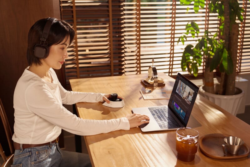 A woman wearing headphones sits at a wooden desk editing video on a laptop. There’s a drink, a wooden tray, and a potted plant nearby. Sunlight streams through wooden blinds in the background.