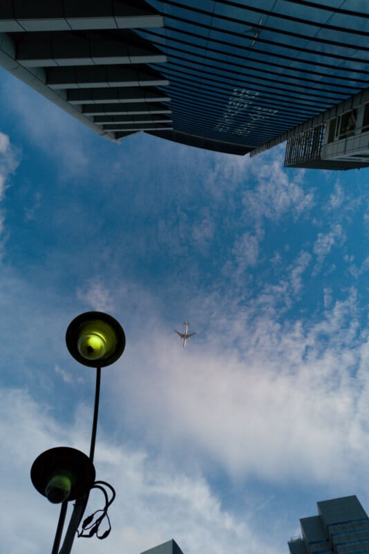 Looking up at a blue sky with scattered clouds, a plane flies overhead between tall modern buildings, while two street lamps with yellow lights are visible in the foreground.