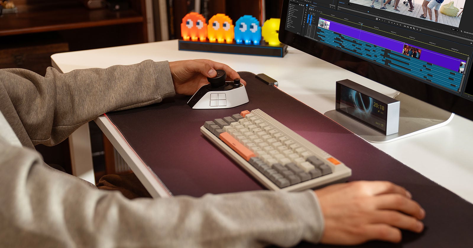 Person sitting at a desk editing video on a computer monitor, using a keyboard and a specialized editing controller. Colorful Pac-Man ghost figures are displayed on the desk in the background.