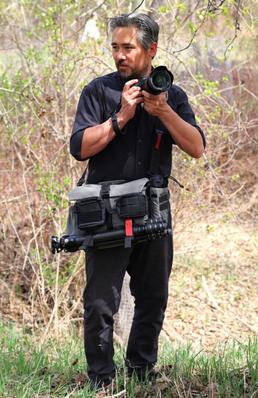 A man with a beard stands outdoors holding a camera. He is dressed in black, carrying a gray camera bag with a tripod attached, surrounded by grass and trees with new leaves.