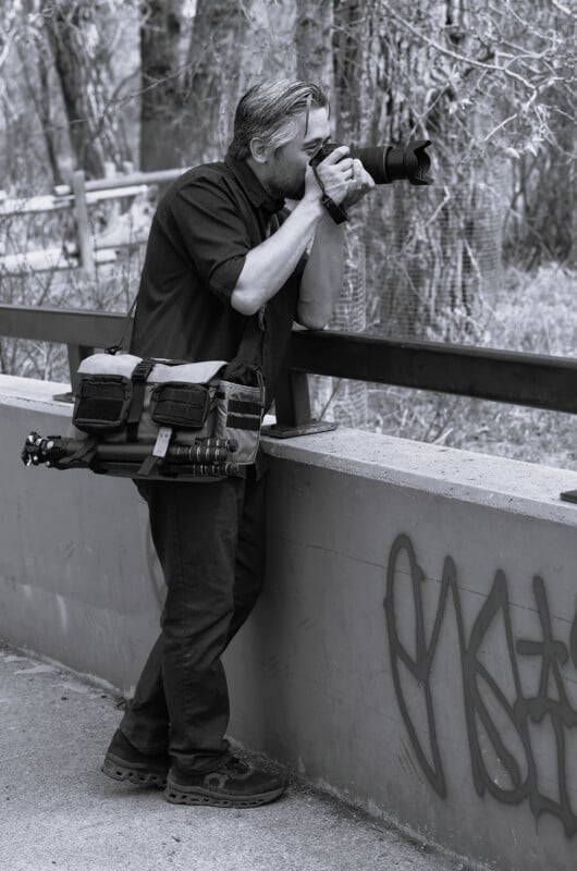 A man stands on a paved path, leaning on a concrete railing with graffiti, photographing something with a DSLR camera and long lens. He has a camera bag and tripod over his shoulder. Trees and a fence are in the background.