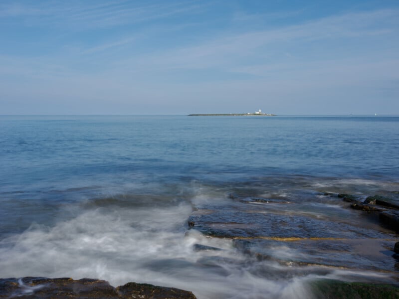 A distant lighthouse sits on a small island under a blue sky, surrounded by calm sea. In the foreground, waves crash gently over dark, rocky shore.