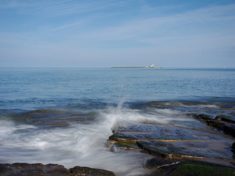 Waves crash onto rocky shore in the foreground, with a calm blue sea stretching to the horizon. An island with a white lighthouse sits in the distance under a clear sky.