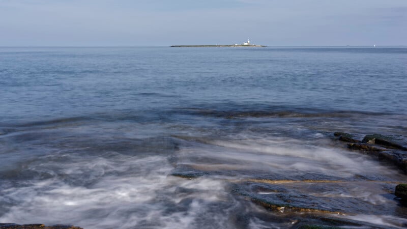 Waves gently wash over rocks in the foreground with calm blue sea stretching to the horizon. In the distance, a small island with a lighthouse is visible under a clear sky.