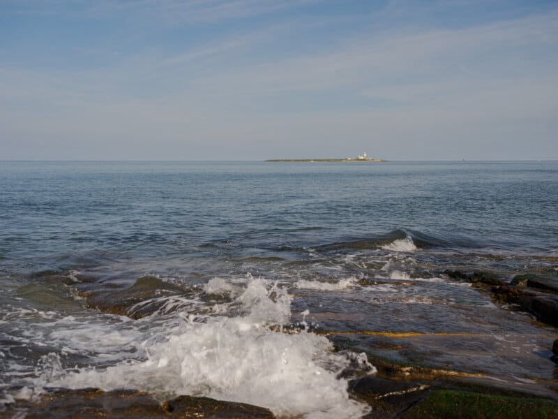 Waves crash onto rocky shore in the foreground, with calm blue sea and a distant island featuring a small white lighthouse under a clear sky.