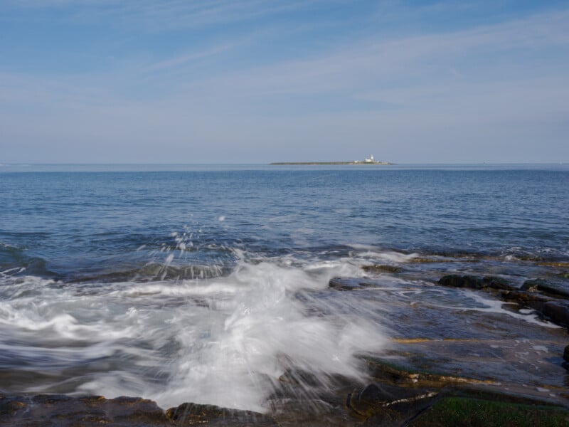 Waves crash against rocky shore in the foreground, with a calm blue sea stretching to the horizon. A small island with a lighthouse is visible in the distance under a lightly clouded sky.
