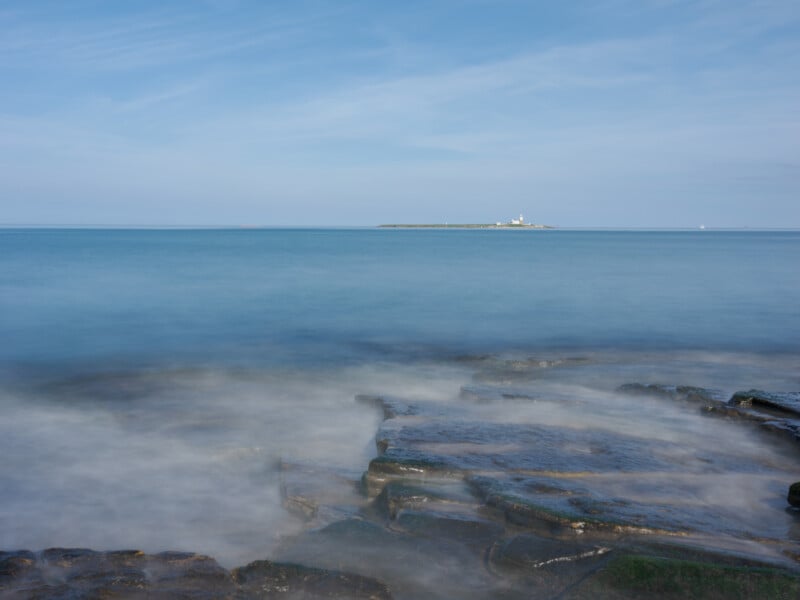 Rocky shoreline with misty water in the foreground, calm blue sea in the midground, and a distant island with a lighthouse under a clear sky in the background.