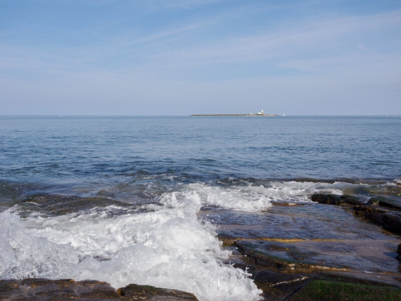 Waves crash onto rocky shore in the foreground, with calm blue sea extending to a small distant island topped with a white lighthouse under a partly cloudy sky.