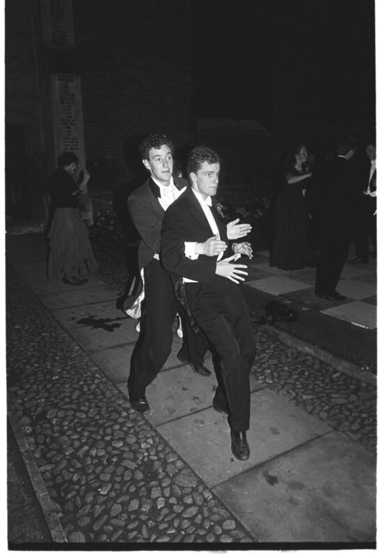 Two men in tuxedos dance playfully in a conga line at an outdoor nighttime event, with other formally dressed people in the background. The setting appears to be a cobblestone walkway near a brick building.