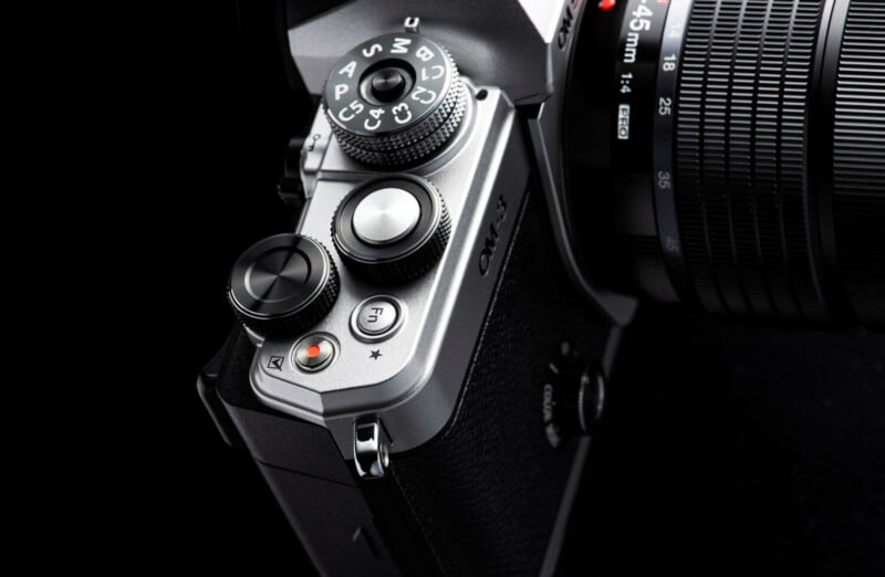 Close-up of the top controls of a silver and black camera, showing dials, buttons, and part of the attached lens against a dark background.