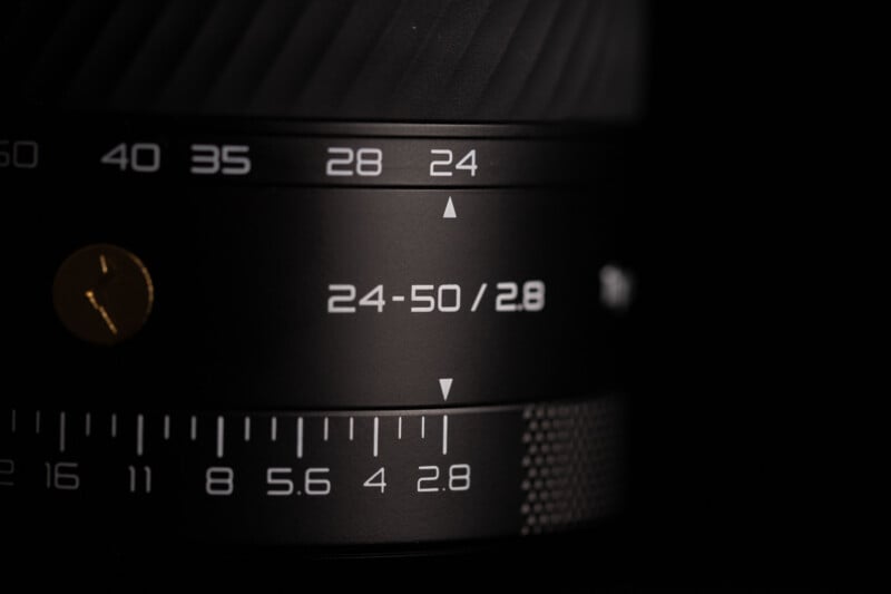 Close-up of a camera lens showing focal length markings (24-50mm) and an aperture value of 2.8, with numbers and arrows indicating settings on the lens barrel.
