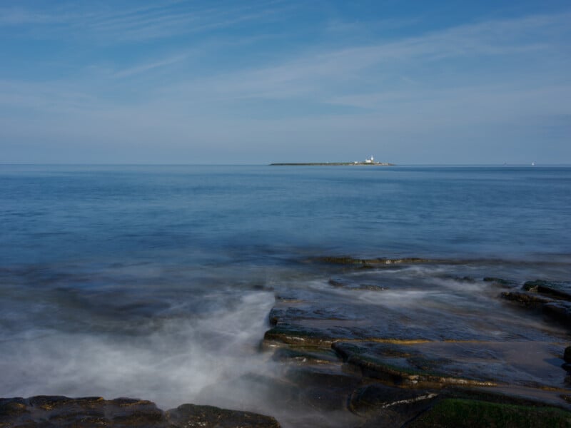 Rocky shoreline with gentle waves in the foreground, calm blue sea, and a distant island with a white lighthouse under a mostly clear sky.