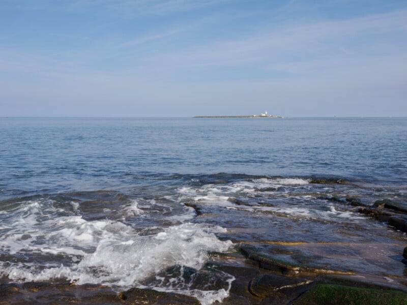 Waves gently crash onto rocky shoreline in the foreground, with calm blue sea stretching to a distant small island featuring a white lighthouse under a pale blue sky.