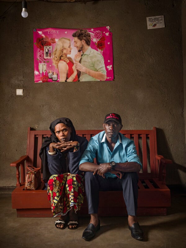 A woman and man sit on a wooden bench in a dim room, both looking serious. Behind them is a bright poster of a romantic couple with the word "LOVE" and pink hearts, contrasting with the plain, brown textured wall.