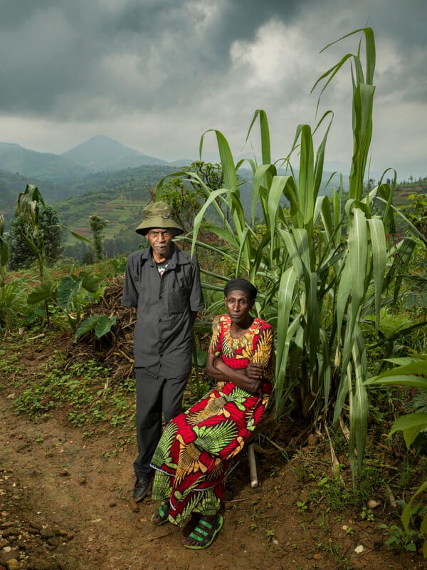 An older man stands beside a seated woman in colorful clothing on a dirt path, with lush green plants and distant hills under a cloudy sky in the background.