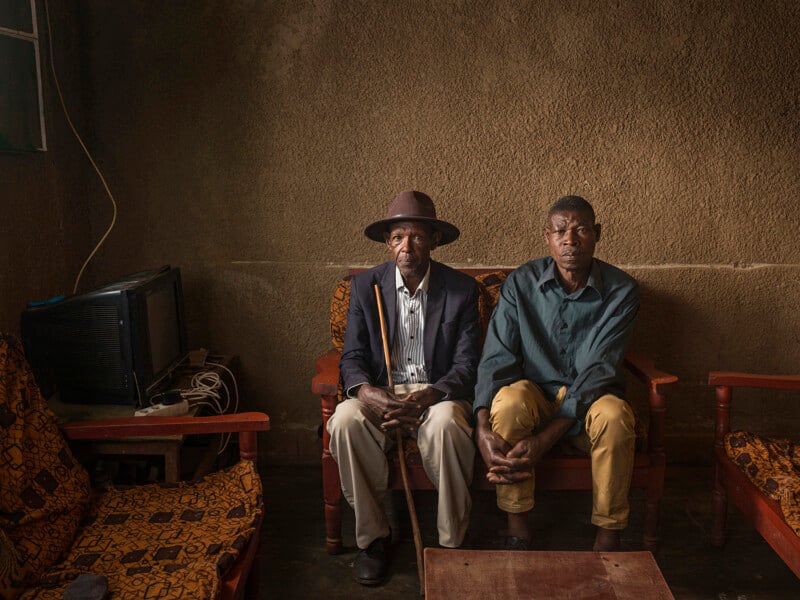 Two men sit side by side on a patterned sofa in a dimly lit room with textured brown walls. One wears a hat and holds a cane; the other wears a green shirt. Old furniture and a TV are in the background.