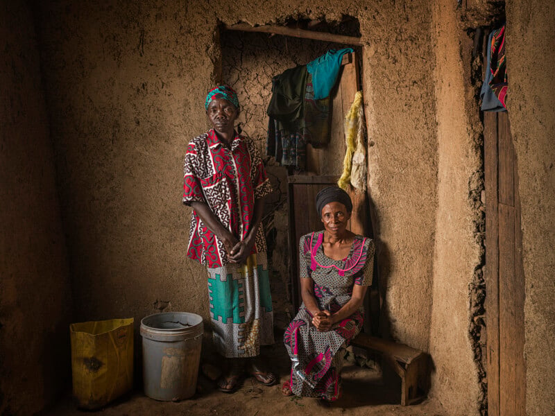 Two women in colorful patterned dresses pose inside a rustic mud-walled room; one stands and the other sits on a bench. A bucket, yellow bag, and hanging clothes are visible in the dimly lit, earthy space.