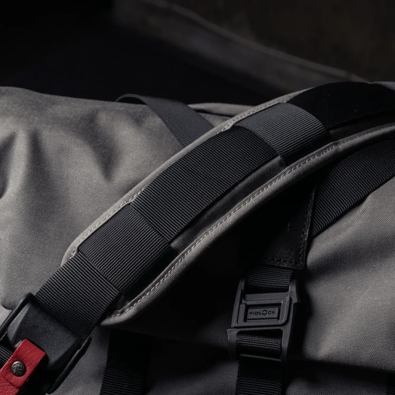 Close-up of a black and gray backpack strap with a padded section, visible stitching, and a plastic buckle, resting on a gray fabric surface.
