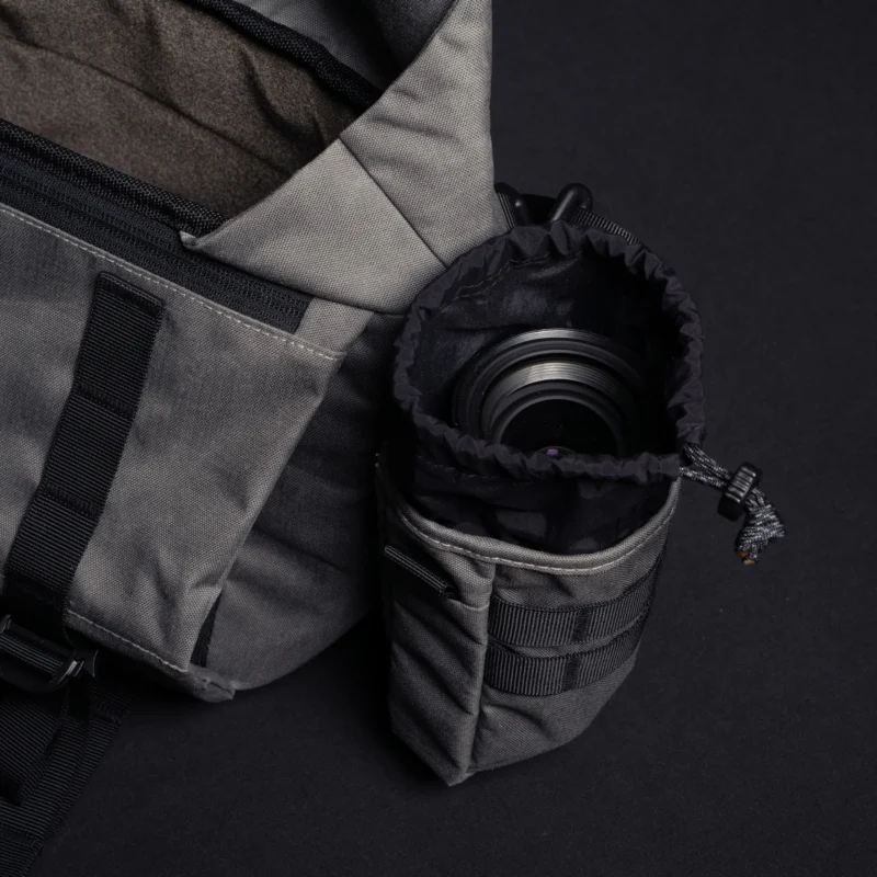 A gray camera bag with a zippered pocket is next to a padded lens pouch holding a camera lens, both sitting on a black surface.