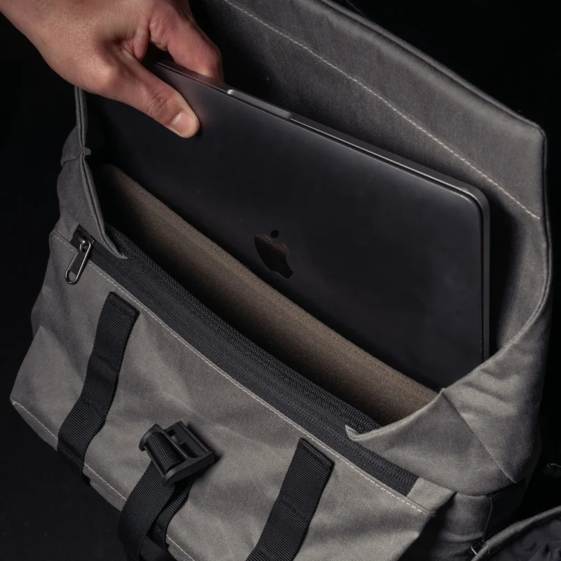 A hand places a closed silver laptop with an Apple logo into a gray fabric messenger bag with black straps and a buckle.