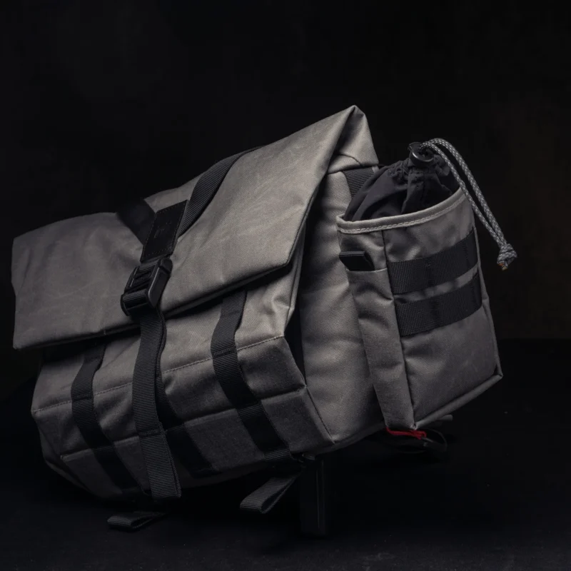 A gray messenger bag with black straps and buckle closures, featuring an external drawstring pocket on the side, displayed against a dark background.