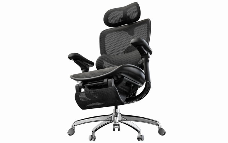 A modern ergonomic office chair with a mesh back, adjustable headrest, armrests, and leg rest, set on a chrome swivel base with five caster wheels. The chair is black with silver accents.