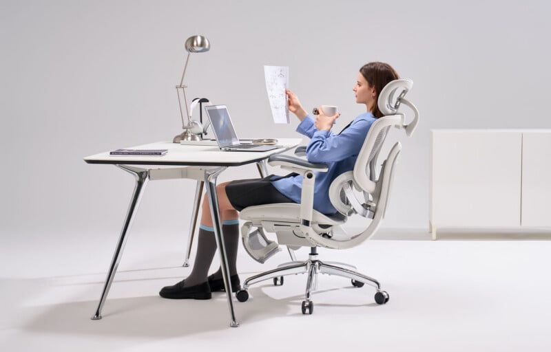 A woman sits in a modern office chair at a minimalist desk, holding a cup and a sheet of paper. The desk has a lamp, a laptop, and notebooks. The background is plain and white.