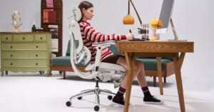A woman sits at a wooden desk, writing on paper, in a modern white office chair. Behind her are a green dresser, a sofa, books, and decorative items. An orange lamp and computer are on the desk.