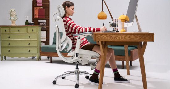 A woman sits at a wooden desk, writing on paper, in a modern white office chair. Behind her are a green dresser, a sofa, books, and decorative items. An orange lamp and computer are on the desk.