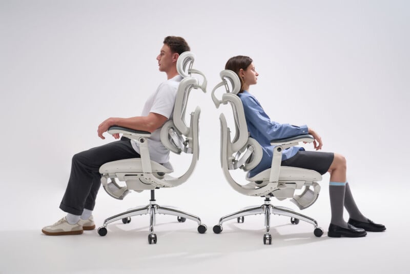 A man and a woman sit back-to-back on white ergonomic office chairs against a plain white background, both facing away from each other with relaxed postures.