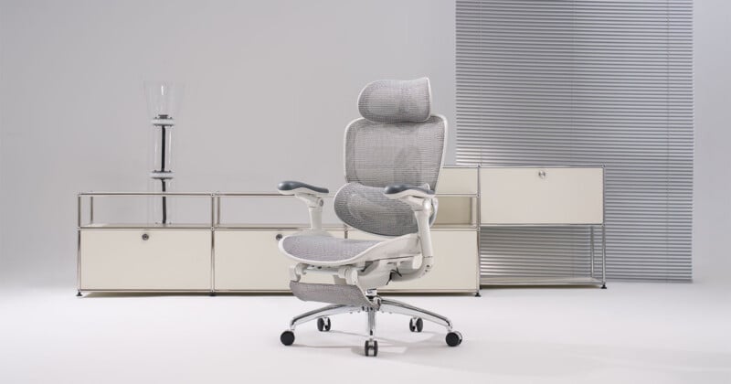 A modern ergonomic office chair with a mesh back and headrest sits in front of minimalist white storage cabinets and a lamp, set against a clean, light-gray background.