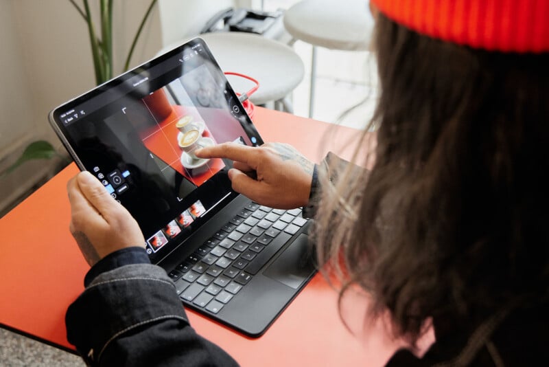 A person wearing a red beanie uses a tablet with a keyboard to edit a photo of two cups of coffee on a red table. The person's finger is touching the screen, adjusting the image.