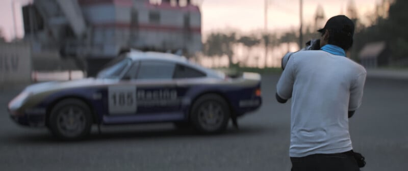 A person in a white shirt and cap takes a photo of a vintage racing car with the number 105, parked on a racetrack with blurred trees and structures in the background.
